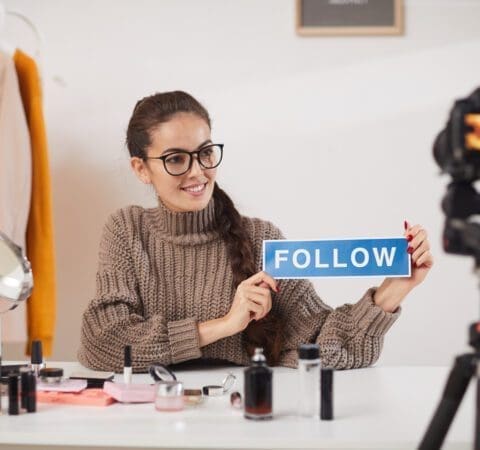 Portrait of contemporary young woman holding FOLLOW word while filming video for beauty and lifestyle channel, copy space