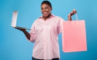 Online shopping. African american woman holding laptop and paper shopper bag standing over blue studio background, smiling to camera. E-commerce, seasonal sales and offers