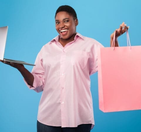 Online shopping. African american woman holding laptop and paper shopper bag standing over blue studio background, smiling to camera. E-commerce, seasonal sales and offers